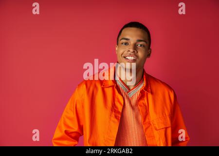bellissimo ragazzo afroamericano con una camicia arancione alla moda, con un sorriso radioso che guarda la fotocamera sul rosso Foto Stock