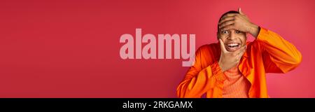 playful african american man looking at camera and touching face, red background, banner, copy space Stock Photo