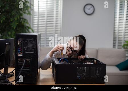 Una giovane donna è seduta in una stanza a una scrivania. Utilizza un cacciavite per rimuovere varie parti dal computer. Il computer è sulla scrivania. In Foto Stock