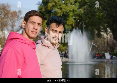 coppia di due giovani della comunità lgbt, che indossano abiti rosa, in piedi in posa guardando la macchina fotografica al tramonto nel parco con una laguna e una fontana d'acqua Foto Stock