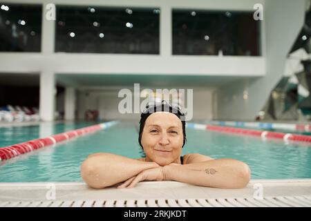 sportive middle aged woman relaxing at poolside, swim cap and goggles, smile, recreation center Foto Stock