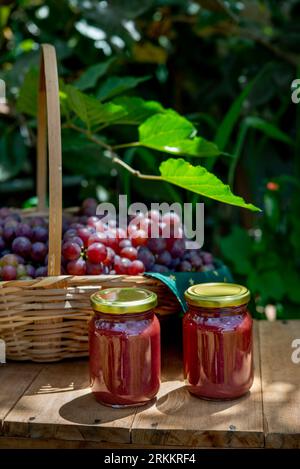 bellissimo cestino pieno di uva fresca con 2 vasetti di marmellata d'uva davanti, vasetti senza labbra Foto Stock