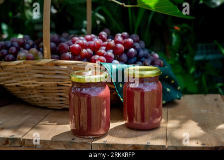 bellissimo cestino pieno di uva fresca con 2 vasetti di marmellata d'uva davanti, vasetti senza labbra Foto Stock