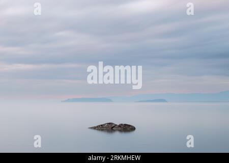 Vista a lunga esposizione di una roccia nel lago Trasimeno dell'Umbria, con isole sullo sfondo e cielo lungoso Foto Stock