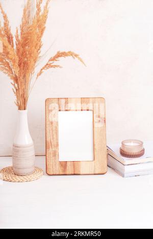 Cornice in legno mockup, libri, candela aromatica e pampas in vaso su tavolo bianco. Copia spazio. Foto Stock