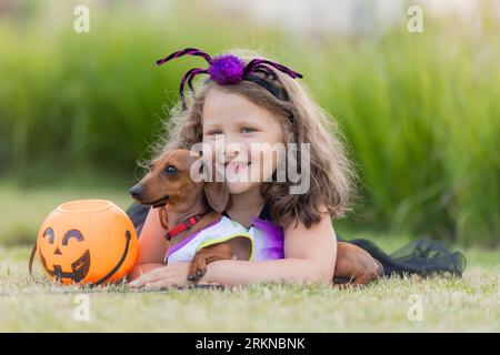 Adorabile bambina in costume da strega per Halloween cammina con un cane da dachshund nel parco. Foto di alta qualità Foto Stock