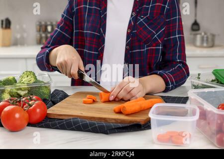 Donna che taglia carote vicino a contenitori con prodotti freschi su un tavolo di marmo bianco in cucina, primo piano. Conservazione degli alimenti Foto Stock