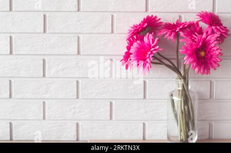 Un bellissimo bouquet di margherite gerbera rosa in un vaso di vetro trasparente si trova sullo sfondo di una parete di mattoni bianchi testurizzati. Foto Stock