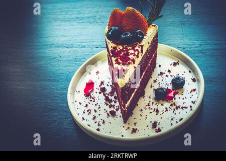 Una fetta gourmet di torta a strati di velluto rosso condita con fragole e mirtilli servita in un ambiente rustico. Foto Stock