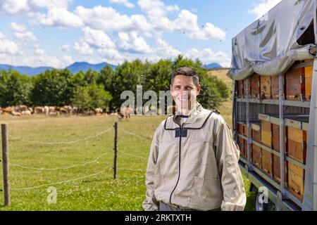 Ritratto di un apicoltore maschio felice che lavora in un apiario vicino ad alveari con api. Foto Stock