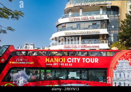 Un autobus rosso a due piani scoperto che offre ai turisti un tour della città di Hanoi passa davanti a diversi famosi caffè alla rotatoria alla fine di Dinh Tien Hoa Foto Stock
