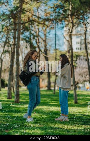 Due ragazze che escono al parco in una giornata di sole. Ragazza dai capelli ricci che guarda qualcosa di divertente al telefono mentre la sua amica sta facendo una telefonata Foto Stock