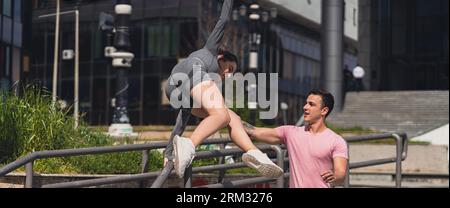 Due amici sportivi che si divertono in città. Una persona maschile in forma che aiuta la sua amica a saltare sopra una recinzione metallica Foto Stock