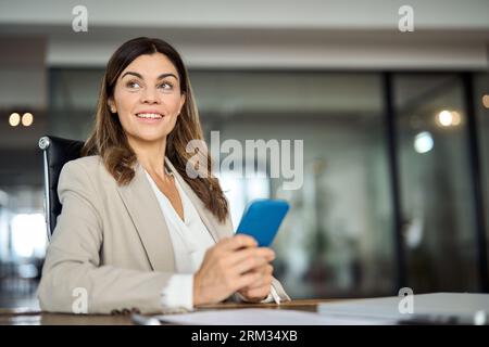 Donna d'affari matura sorridente che tiene il telefono e guarda lontano in ufficio. Foto Stock