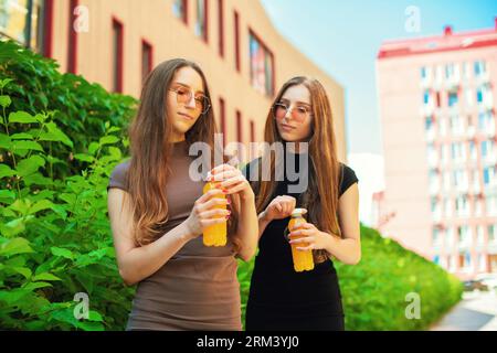 Giovani sorelle gemelle urbane con occhiali da sole freschi che aprono una bottiglia di succo di limone sullo sfondo della strada della città Foto Stock