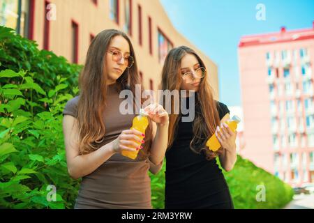 Giovani sorelle gemelle urbane con occhiali da sole freschi che aprono una bottiglia di succo di limone sullo sfondo della strada della città Foto Stock