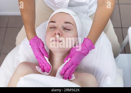 Il cosmetologo Top View pulisce la pelle del collo e del viso della giovane donna prima della procedura di bellezza, ringiovanimento nel centro benessere. Paziente che riceve ago Foto Stock