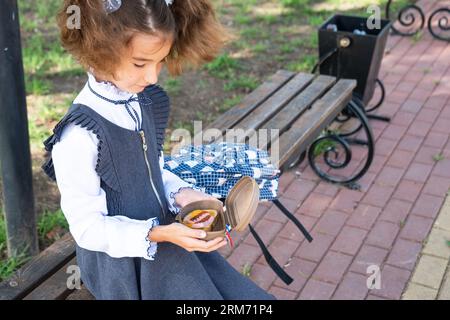 Ragazza con zaino che mangia panino confezionata in una scatola vicino a scuola. Uno spuntino veloce con un panino, cibo malsano, pranzo da scuola. Torna a scuola. Foto Stock