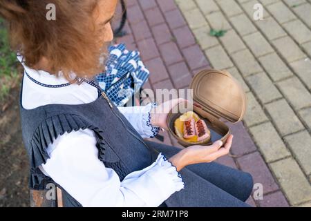 Ragazza con zaino che mangia panino confezionata in una scatola vicino a scuola. Uno spuntino veloce con un panino, cibo malsano, pranzo da scuola. Torna a scuola. Foto Stock
