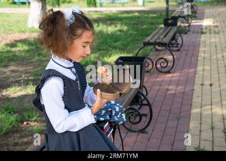 Ragazza con zaino che mangia panino confezionata in una scatola vicino a scuola. Uno spuntino veloce con un panino, cibo malsano, pranzo da scuola. Torna a scuola. Foto Stock