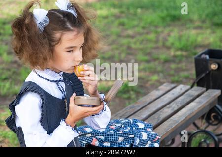 Ragazza con zaino che mangia panino confezionata in una scatola vicino a scuola. Uno spuntino veloce con un panino, cibo malsano, pranzo da scuola. Torna a scuola. Foto Stock