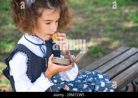 Ragazza con zaino che mangia panino confezionata in una scatola vicino a scuola. Uno spuntino veloce con un panino, cibo malsano, pranzo da scuola. Torna a scuola. Foto Stock
