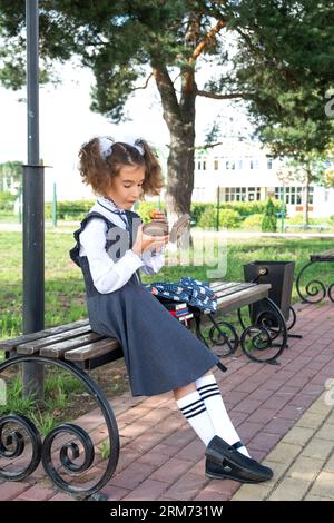 Ragazza con zaino che mangia panino confezionata in una scatola vicino a scuola. Uno spuntino veloce con un panino, cibo malsano, pranzo da scuola. Torna a scuola. Foto Stock