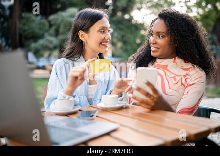 Due amiche Happy Female che usano smartphone e carta di credito al Cafe Outdoors Foto Stock