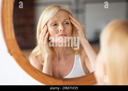 Donna di mezza età sconvolta che guarda lo specchio e tocca le rughe sul viso, donna che esamina le linee sottili sulla fronte Foto Stock
