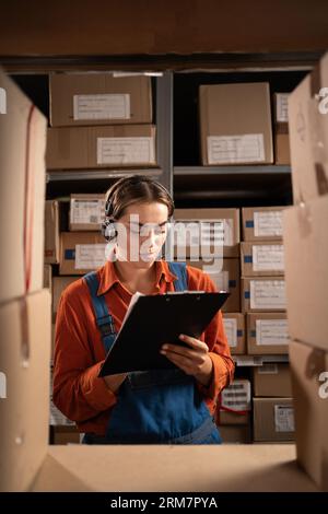 Operatore professionale femminile che indossa cuffie in piedi accanto allo scaffale, conta i pacchi nel magazzino e fa inventario delle merci Foto Stock