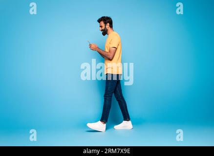 Ragazzo indiano in abito casual che cammina con smartphone e sorride su sfondo blu studio, a tutta lunghezza, vista laterale Foto Stock