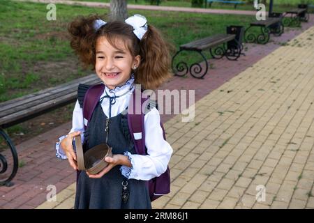 Ragazza con zaino che mangia panino confezionata in una scatola vicino a scuola. Uno spuntino veloce con un panino, cibo malsano, pranzo da scuola. Torna a scuola. Foto Stock
