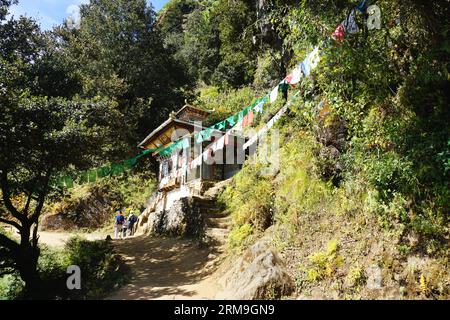 Gli escursionisti camminano davanti a un tradizionale edificio bhutanese con bandiere di preghiera sopra il sentiero per il Monastero del Nido della Tigre (Taktsang) vicino a Paro, in Bhutan Foto Stock