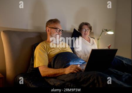 Coppia anziana che usa un computer portatile sdraiato sul letto in camera da letto a casa confortevole Foto Stock