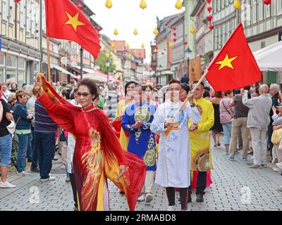 27 agosto 2023, Sassonia-Anhalt, Wernigerode: Partecipanti a una marcia di sfilata attraverso il centro storico di Wernigerode con una bandiera nazionale della repubblica socialista del Vietnam. Con il festival delle lanterne a Wernigerode, il decimo anniversario del gemellaggio tra Hoi An e Wernigerode è debitamente celebrato. La città vecchia di Wernigerode risplende di nuovo alla luce di centinaia di lanterne vietnamite fatte a mano. La città portuale vietnamita di Hoi An è stata la città gemella di Wernigerode dall'agosto 2013. La città, con circa 75.000 abitanti, si trova sulla costa del Vietnam centrale sul Mar Cinese meridionale, circa 30 k Foto Stock