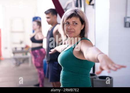 Donna caucasica di mezza età con i capelli biondi e gli occhi blu in una palestra che si esercita con i colleghi. Foto Stock