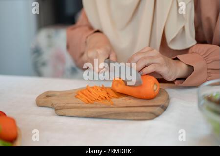 Immagine ravvicinata di una donna musulmana che prepara gli ingredienti per il suo mix di insalate in cucina, affetta la carota con il coltello su un tagliere, cucina in th Foto Stock