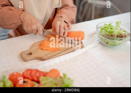 Immagine ravvicinata di una donna musulmana che prepara gli ingredienti per il suo mix di insalate in cucina, affetta la carota con il coltello su un tagliere, cucina in th Foto Stock