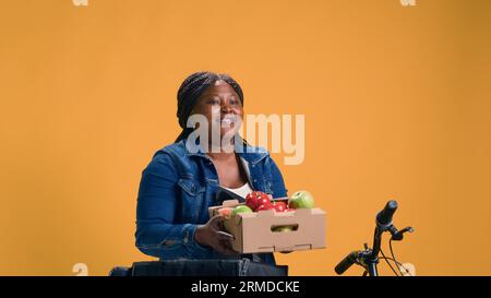 Cestino di frutta consegnato delicatamente al sacchetto per la consegna del cibo da parte di un messaggero afro-americano. Corriere afro-americano in bicicletta mentre sorseggia mentre consegna prodotti freschi. Foto Stock