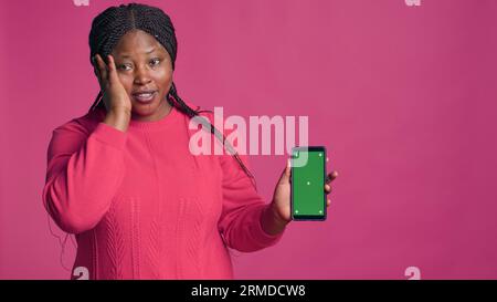 La bellezza afro-americana femminile tiene verticalmente il dispositivo mobile che visualizza uno schermo verde. Splendida donna nera che mostra un'innovativa app per lo shopping online sullo smartphone con modello di spazio copioso vuoto. Foto Stock