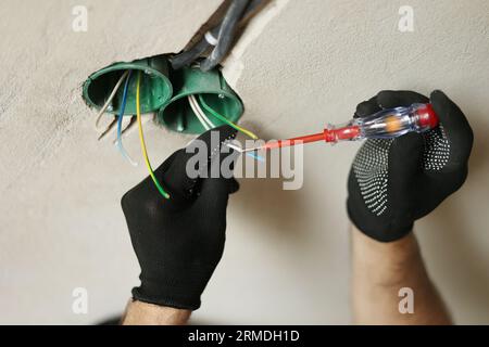 Il maschio per fotoelettricista ritagliato in guanti protettivi controlla la presenza di tensione elettrica nella fase della presa. Controllo dei fili di tensione nella presa utilizzando Foto Stock