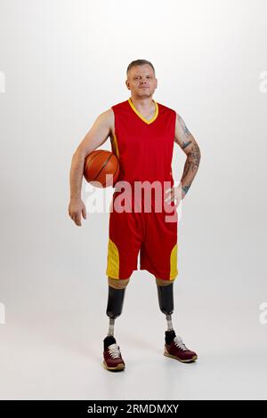 Attraente amputato uomo con protesi alle gambe invalidità in piedi che regge la palla da basket arancione. Sport inclusivo per disabili. Foto Stock