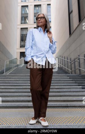 ritratto aziendale di una donna anziana di successo con capelli grigi e occhiali vestiti con abiti eleganti da ufficio sullo sfondo della città Foto Stock