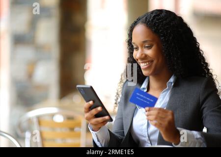 Felice donna d'affari nera che compra online con carta di credito e smartphone in un bar sulla terrazza Foto Stock