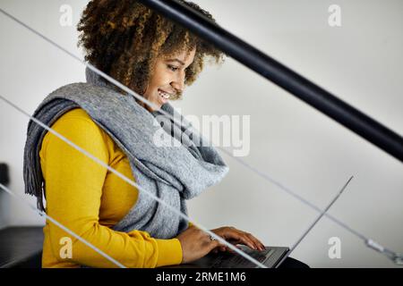 Vista laterale di una donna d'affari sorridente con i capelli ricci che utilizza un computer portatile mentre è seduta sui gradini dell'ufficio Foto Stock