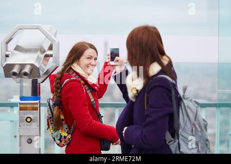 Due ragazze allegri a Parigi, che si fotografano l'una sull'altra sulla piattaforma di osservazione della torre di Montparnasse con il cellulare Foto Stock