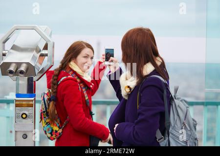 Due ragazze allegri a Parigi, che si fotografano l'una sull'altra sulla piattaforma di osservazione della torre di Montparnasse con il cellulare Foto Stock