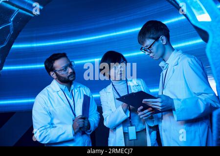 scienziati multietnici che guardano giovani stagisti con clipboard in un centro scientifico futuristico illuminato al neon Foto Stock