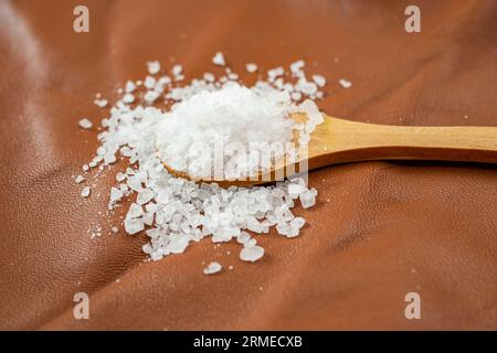 sale di roccia bianca che riempie un cucchiaino di legno, su pelle marrone, texture della pelle, vista laterale, sfondo astratto con messa a fuoco morbida Foto Stock
