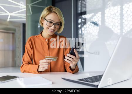 Giovane bella donna d'affari all'interno dell'ufficio sul posto di lavoro, con in mano un telefono e una carta di credito, una dipendente seduta al tavolo che effettua acquisti online e trasferimenti di denaro. Foto Stock